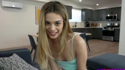 Blonde Schönheit mit langen Haaren lächelt verführerisch in hellblauem Top in modernem Wohnzimmer