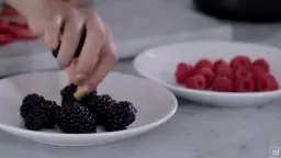 Hand legt eine Himbeere auf frische Brombeeren in einer weißen Schale in der Küche