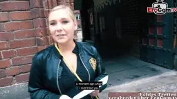 Blonde Frau mit offenem Lederblouson zeigt ihre großen nackten Brüste vor roter Backsteinwand